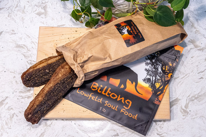 Two strips of dark dried meat in a brown paper bag on a wood board next to a Lowfeld Soul Food Biltong 1 lb package.