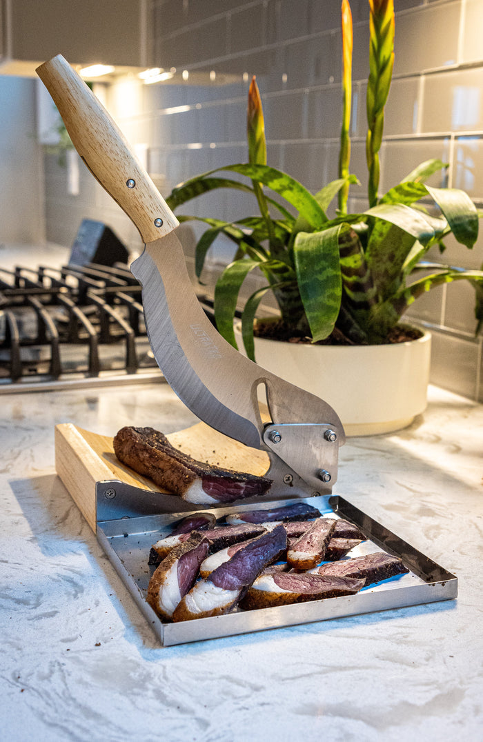 Slices of dried meat on a metal tray under a wood-handled biltong slicer on a marble countertop.