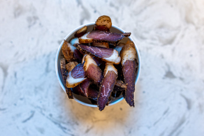Slices of dark, seasoned biltong meat with white fat edges in a small white ceramic bowl on a light stone surface.
