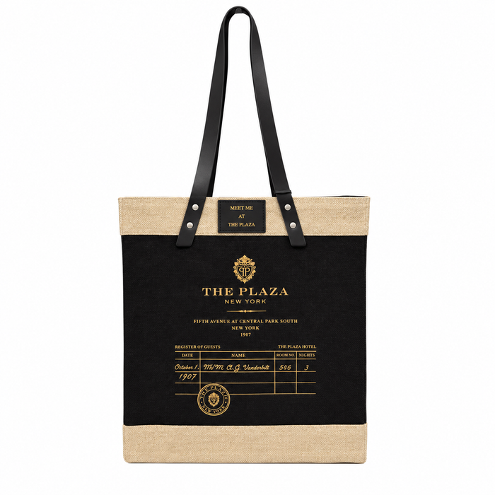 A tote bag styled like a vintage hotel check-in slip, featuring a black central panel with gold text reading 'THE PLAZA NEW YORK' and a beige jute top and bottom trim, with black shoulder straps.